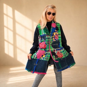 May include: A sleeveless waistcoat featuring a patchwork design of green plaid fabric and dark blue denim. The garment is embellished with various floral fabric appliques, pink and blue lace trim, and frayed edges, worn over a black long-sleeve shirt.