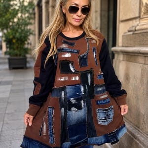 Puede incluir: Una sudadera marrón de manga larga con un diseño de patchwork hecho de varios retazos de tela de mezclilla azul, incluyendo bolsillos y bordes deshilachados, superpuestos en el frente. Las mangas son de color negro sólido y tiene cuello redondo.