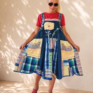 Puede incluir: Un vestido tipo pichi de mezclilla sin mangas con un diseño de patchwork de paneles de tela a cuadros azules, verdes y amarillos, dos bolsillos amarillos grandes con borde de encaje y una aplicación floral amarilla central, sobre una camiseta roja.