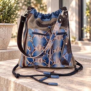 Peut inclure: Un sac à bandoulière multi-texturé avec un panneau central à motif peau de serpent bleu et marron, des panneaux latéraux en daim beige et des détails en cuir marron foncé. Le sac comprend une fermeture à cordon, une poche zippée et des lanières noires.