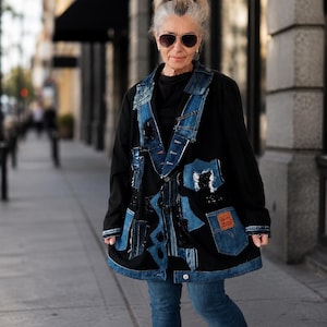 May include: A black jacket with denim patches and accents. The jacket features a patchwork design with various shades of blue denim and decorative elements. The person is wearing blue jeans and sunglasses.