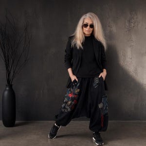 以下が含まれることがあります： 黒のブレザー、黒のトップス、右脚にカラフルな花柄の刺繍、左脚にパッチワークのディテールが施された黒のハーレムパンツ、黒のスポーツシューズ。ダークサングラスをかけ、長いブロンドヘアのスタイル。