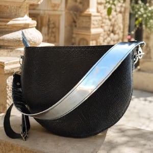 May include: A black, semi-circular shoulder bag featuring a textured, reptilian-like pattern on its surface. The bag is equipped with a wide, reflective silver strap that contrasts with the dark body, attached via silver-toned metal hardware.
