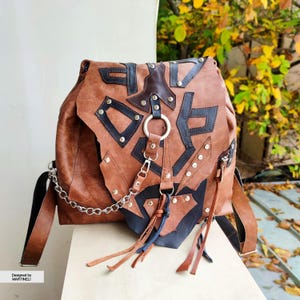 May include: Brown leather backpack with black geometric accents, silver studs, and a silver chain strap. Features a large metal ring and decorative leather tassels. The backpack is designed by MARTINELLI.