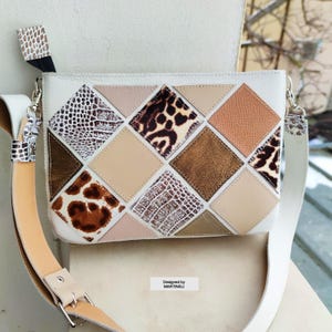 May include: White crossbody bag with a geometric design featuring various textures and patterns, including animal prints and metallic accents. The bag has a long, adjustable strap and a smaller strap with a matching print. The text "Designed by MARTINELLI" is visible.