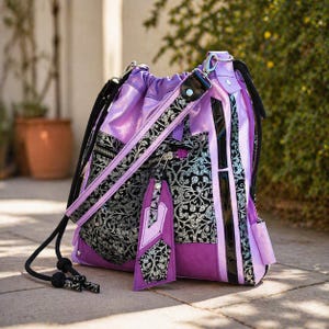 May include: A purple and black drawstring backpack with a decorative floral pattern. The bag features a black strap, a zipper, and a matching tag. The bag is made of a shiny, textured material.