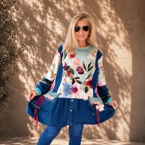 Könnte beinhalten: Eine langärmelige Tunika mit hellblauem Blumenmuster am Oberteil und an den Ärmeln, kontrastiert durch einen unteren Teil aus dunkelblauem Denim. Das Kleidungsstück verfügt über zwei große Vordertaschen mit floralen Akzenten und roten Bändern.