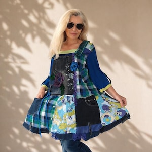 Op de afbeelding: Een patchwork tuniek bestaande uit een mix van blauwe denim, groen en blauw geruite stof, en een bloemenprint in tinten blauw, groen en wit. Het kledingstuk bevat meerdere denim zakken, decoratieve stoffen bloemen en lange mouwen.