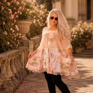 Op de afbeelding: Een perzikkleurige tuniek met een doorschijnend bovenlijfje, lange mouwen en een uitlopende rok met gemengde patronen van bloemen en abstracte vormen. Het kledingstuk heeft twee voorzakken versierd met pailletten en een kanten zoom.