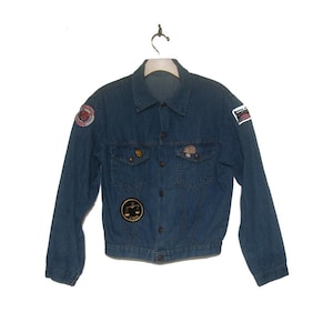 May include: A blue denim jacket with multiple patches, including a scale of justice, a mushroom, and a red and white patch. The jacket has a button-up front and long sleeves.
