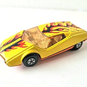 May include: Yellow toy car with black and red flames painted on the hood. The car has a clear windshield and black wheels.