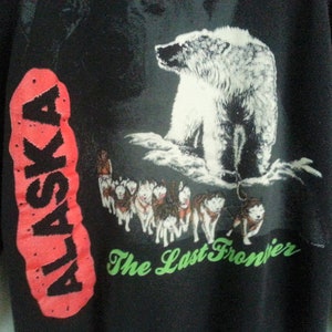 May include: Black sweatshirt with a red and black "ALASKA" graphic on the left sleeve. The right side of the sweatshirt features a white polar bear standing on a snowy mountain with a group of sled dogs running below. The text "The Last Frontier" is printed in green below the image.