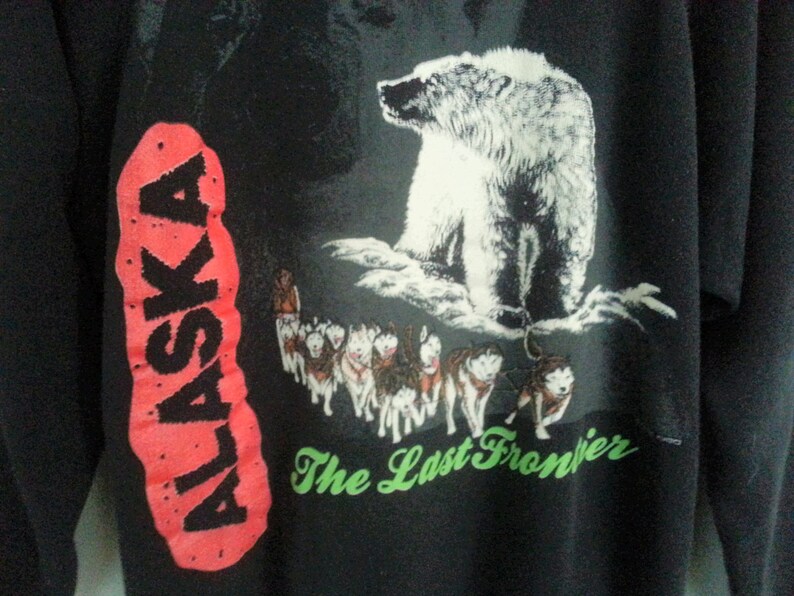 May include: Black sweatshirt with a red and black "ALASKA" graphic on the left sleeve. The right side of the sweatshirt features a white polar bear standing on a snowy mountain with a group of sled dogs running below. The text "The Last Frontier" is printed in green below the image.