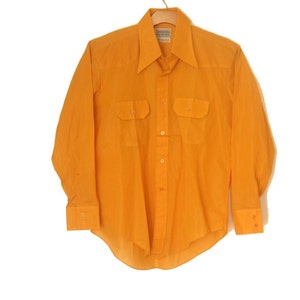May include: Bright orange long-sleeved shirt with two chest pockets and a pointed collar. The shirt is button-down and hangs on a wooden hanger. The cuffs have button closures. The shirt is a solid color.