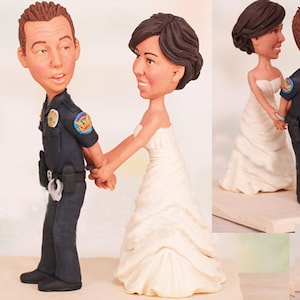 May include: A wedding cake topper featuring a bride and groom figurine. The groom is dressed in a police uniform with a badge and handcuffs. The bride is wearing a white wedding dress. The figurines are made of painted clay.