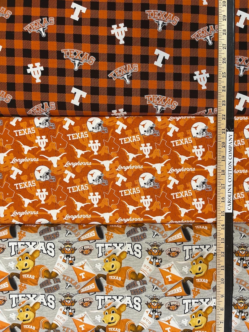 K&ouml;nnte beinhalten: Drei verschiedene Stoffe mit Texas-Motiv. Der obere Stoff ist orange und schwarz kariert mit "Texas"- und "T"-Logos. Der mittlere Stoff ist orange mit "Texas", Helmen und Longhorn-Logos. Der untere Stoff ist grau mit dem Texas-Maskottchen.