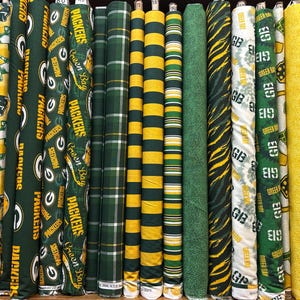 May include: A collection of fabric bolts featuring Green Bay Packers team branding, including logos, text, and team colors of green and gold. The patterns include stripes, plaids, solid yellow, and tie-dye designs, all arranged vertically in a row.