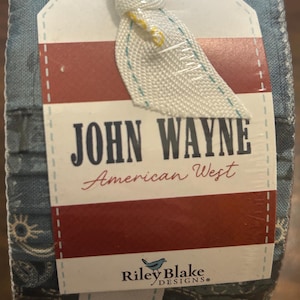 May include: A bundle of fabric with a tag featuring the text "JOHN WAYNE American West." The tag has a red and white striped design and the Riley Blake Designs logo. The fabric has a blue and white pattern.