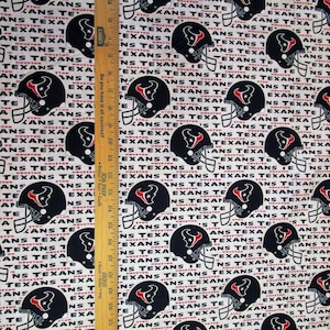 May include: Fabric featuring a repeating pattern of black Houston Texans football helmets with the team name in white and red on a white background. A wooden ruler is in the image.