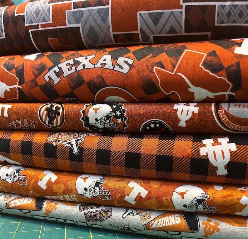K&ouml;nnte beinhalten: Ein Stapel gemusterter Stoffrollen in Orange, Schwarz und Grau. Designs beinhalten das Wort "TEXAS", die Umrisse des Staates, Footballhelme und das Wort "LONGHORNS". Der Stoff hat ein College-Thema.