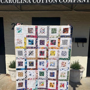 May include: A patchwork quilt featuring a grid of square fabric panels with diverse graphic prints, including sports team logos, checkered patterns, and various colorful motifs, all framed by white fabric borders and finished with a light-colored trim.