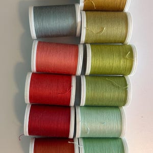 May include: A collection of spools of thread in various colors, including brown, gold, teal, red, and green. The spools are arranged in rows, with white and black accents. The thread appears to be for sewing or crafting projects.