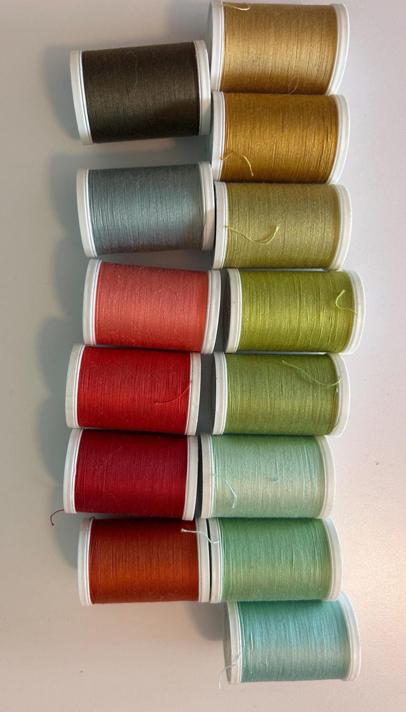 May include: A collection of spools of thread in various colors, including brown, gold, teal, red, and green. The spools are arranged in rows, with white and black accents. The thread appears to be for sewing or crafting projects.