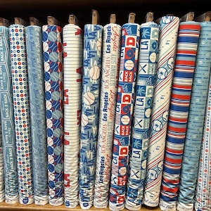 May include: A collection of fabric rolls with baseball-themed designs. The fabrics feature patterns with baseballs, team logos, and text, in shades of blue, red, and white. Some rolls have the text "Los Angeles" and "Back 2 Back".
