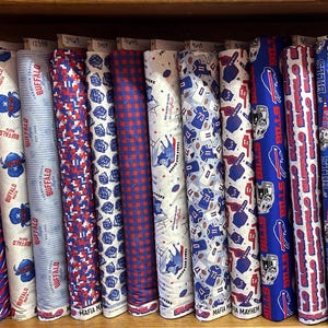 May include: A collection of twelve fabric bolts arranged on a wooden shelf, featuring various patterns in red, white, and blue. The designs include Buffalo Bills team logos, football jerseys, helmets, and text such as Buffalo Mafia and Bills.