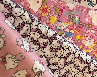Op de afbeelding: Drie gevouwen stukken stof met Hello Kitty-patronen. Eén stof is wit met regenbogen en Hello Kitty, één is roze met bloemen en Hello Kitty, en één is roze met een herhalend patroon van Hello Kitty in een tuinbroek.