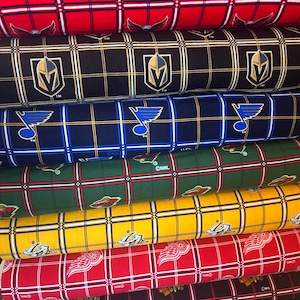 May include: A stack of fabric rolls featuring various NHL team logos and plaid patterns. The fabrics display logos for teams like the Washington Capitals, Vegas Golden Knights, and Detroit Red Wings. Colors include red, black, blue, green, and yellow.