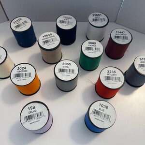 May include: A collection of spools of thread in various colors, including navy, black, white, tan, green, red, gray, orange, purple, and blue. Each spool has a label with a unique number and color name. The thread is likely for sewing or crafting.