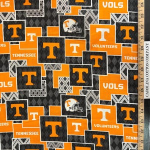 May include: Fabric with a repeating pattern of orange and white "T" logos, football helmets, and the words "VOLS" and "TENNESSEE" on a black and gray diamond background. The fabric is likely cotton.