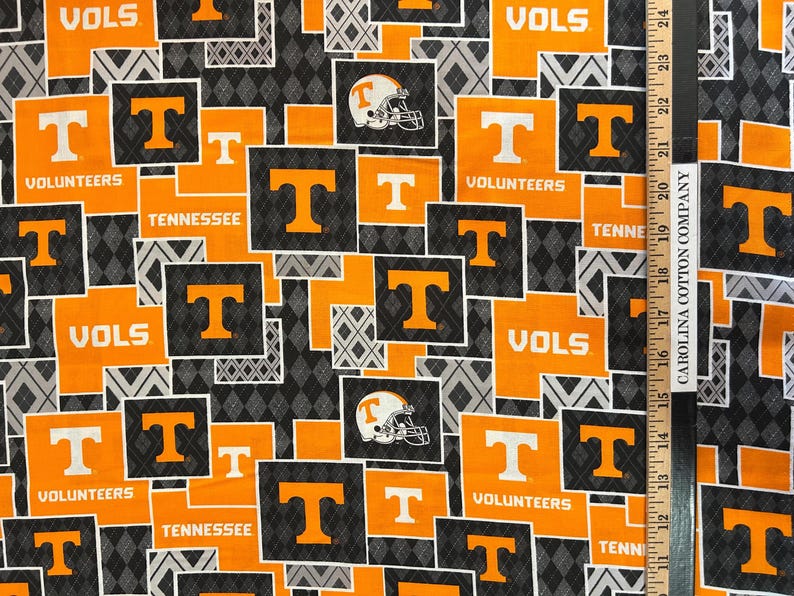 May include: Fabric with a repeating pattern of orange and white "T" logos, football helmets, and the words "VOLS" and "TENNESSEE" on a black and gray diamond background. The fabric is likely cotton.