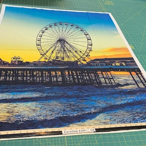 Peut inclure: Un tissu imprimé coloré représentant une grande roue sur une jetée surplombant une mer bleue avec des couleurs de coucher de soleil jaune et orange en arrière-plan.