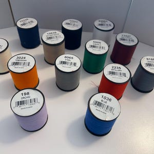 May include: A collection of colorful spools of thread, each with a label displaying a unique color name and number. Colors include blue, orange, green, red, and gray. The spools are arranged on a white surface, ideal for sewing and crafting projects.