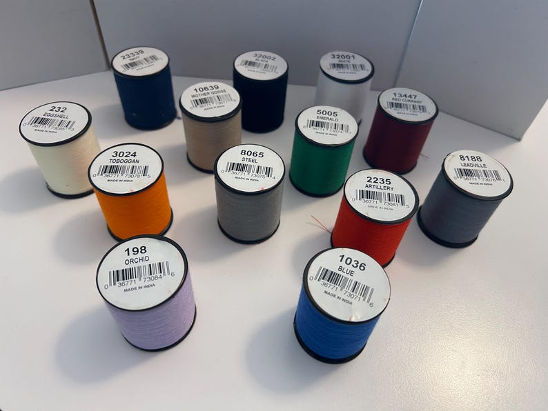 May include: A collection of colorful spools of thread, each with a label displaying a unique color name and number. Colors include blue, orange, green, red, and gray. The spools are arranged on a white surface, ideal for sewing and crafting projects.