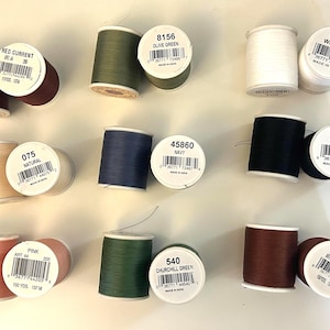 Könnte beinhalten: Eine Sammlung von acht Nähgarnrollen in verschiedenen Farben, darunter Johannisbeerrot, Olivgrün, Weiß, Natur, Marineblau, Schwarz, Pink, Churchill-Grün und Rost. Jede Rolle hat ein weißes Etikett mit Farbnamen, Codes und Barcodes.
