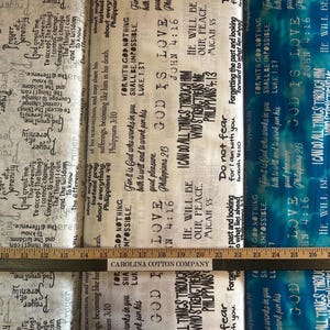 May include: Fabric with various religious text prints in black and gray on a white background, with a blue and white fabric section. A wooden ruler is placed across the fabric.