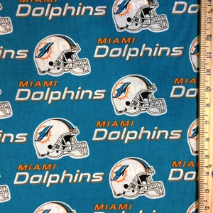 May include: Teal fabric featuring a repeating pattern of Miami Dolphins football helmets and the team name in white and orange. The helmets have a white base with an orange and teal logo. A wooden ruler is visible on the right side.