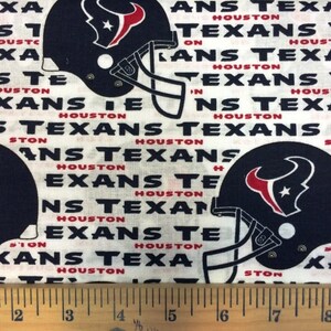 May include: White fabric with a repeating pattern of navy blue Houston Texans football helmets and the words "TEXANS" and "HOUSTON" in black and red. The fabric is likely cotton or a similar material.