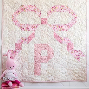 May include: A handmade baby quilt with a large pink bow design and the letter "P" in the center. The quilt is cream-colored with a pink floral pattern on the bow and a pink border. A stuffed bunny sits beside the quilt.