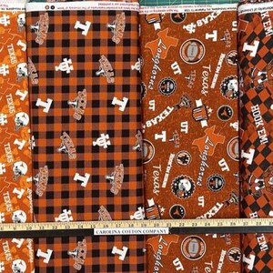 K&ouml;nnte beinhalten: Eine Auswahl an Stoffmustern mit dem Text "TEXAS" und Longhorn-Motiven in Orange, Wei&szlig; und Schwarz. Designs umfassen sich wiederholende Muster, Karomuster und Logoanordnungen. Ein Lineal liegt unten.