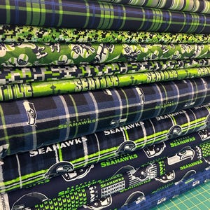 May include: A stack of folded fabric bolts featuring Seattle Seahawks themed patterns in navy blue, lime green, and white. Designs include plaid, digital camouflage, football player silhouettes, the team logo, and the text Seattle Seahawks.