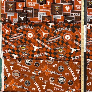K&ouml;nnte beinhalten: Stapel von Stoffen mit verschiedenen Texas Longhorns-Designs. Die Muster umfassen den Umriss des Bundesstaates Texas, die W&ouml;rter "Texas" und "Longhorns" sowie den Satz "Hook 'Em Horns". Das Farbschema ist Orange, Wei&szlig; und Schwarz.