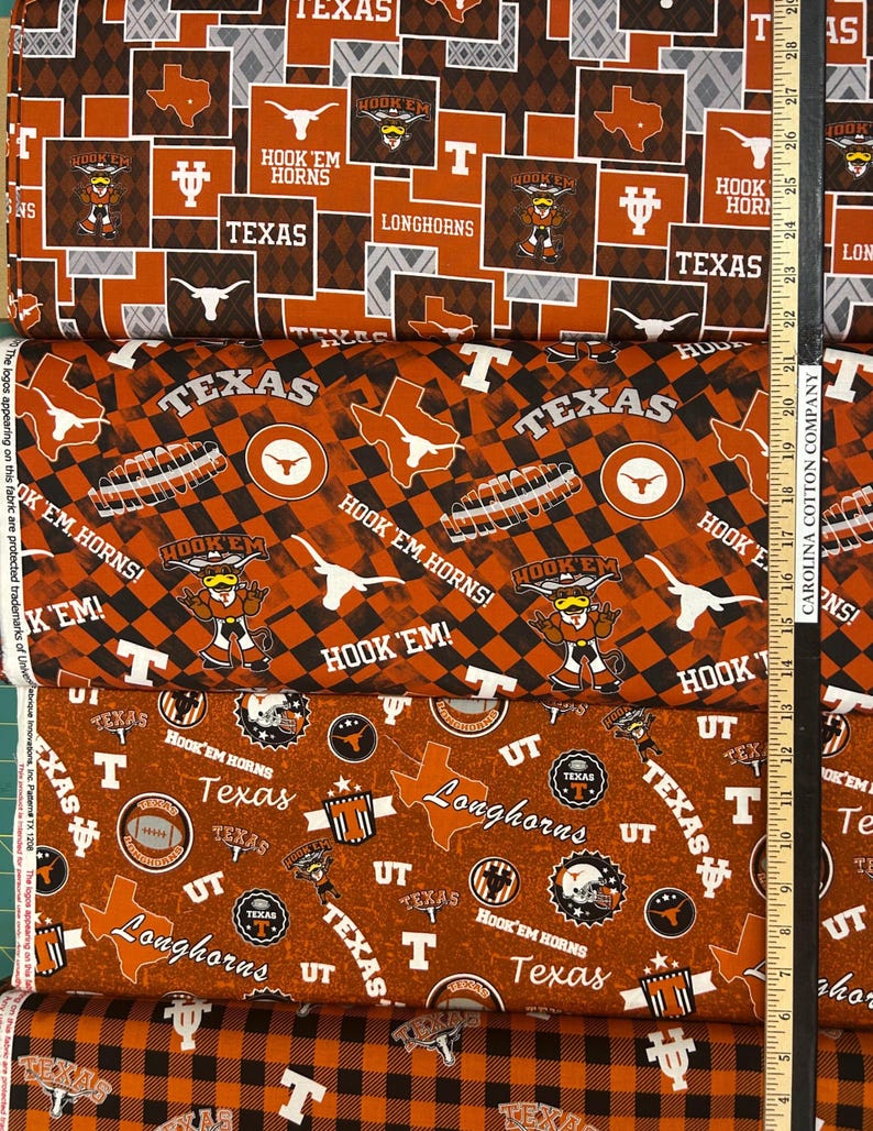 K&ouml;nnte beinhalten: Stapel von Stoffen mit verschiedenen Texas Longhorns-Designs. Die Muster umfassen den Umriss des Bundesstaates Texas, die W&ouml;rter "Texas" und "Longhorns" sowie den Satz "Hook 'Em Horns". Das Farbschema ist Orange, Wei&szlig; und Schwarz.