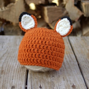May include: A crocheted orange fox hat with white and black ears. The hat is lying on a wooden surface.