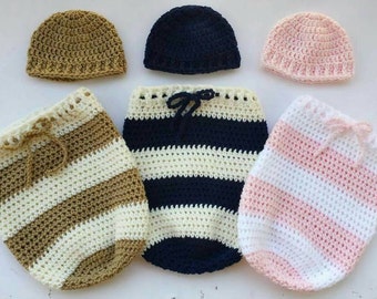 May include: Three crocheted baby hats and three crocheted baby sleeping sacks. The hats are brown, navy blue, and pink. The sleeping sacks are brown and white striped, navy blue and white striped, and pink and white striped.