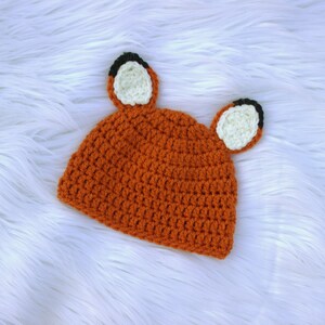 May include: A crocheted orange fox hat with black and white ears. The hat is on a white fluffy surface.