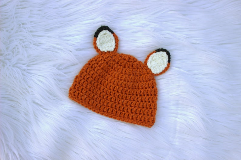 May include: A crocheted orange fox hat with black and white ears. The hat is on a white fluffy surface.